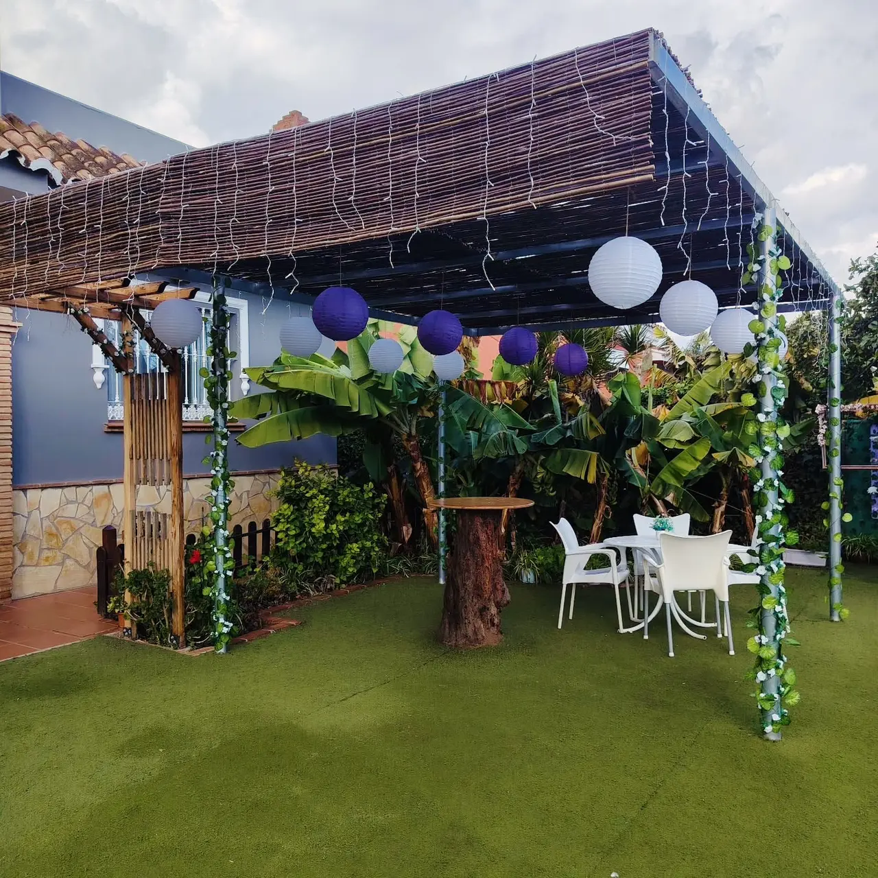 Pérgola chill-out con techo de cañizo, farolillos y plataneras junto a la casa de Villa Meli